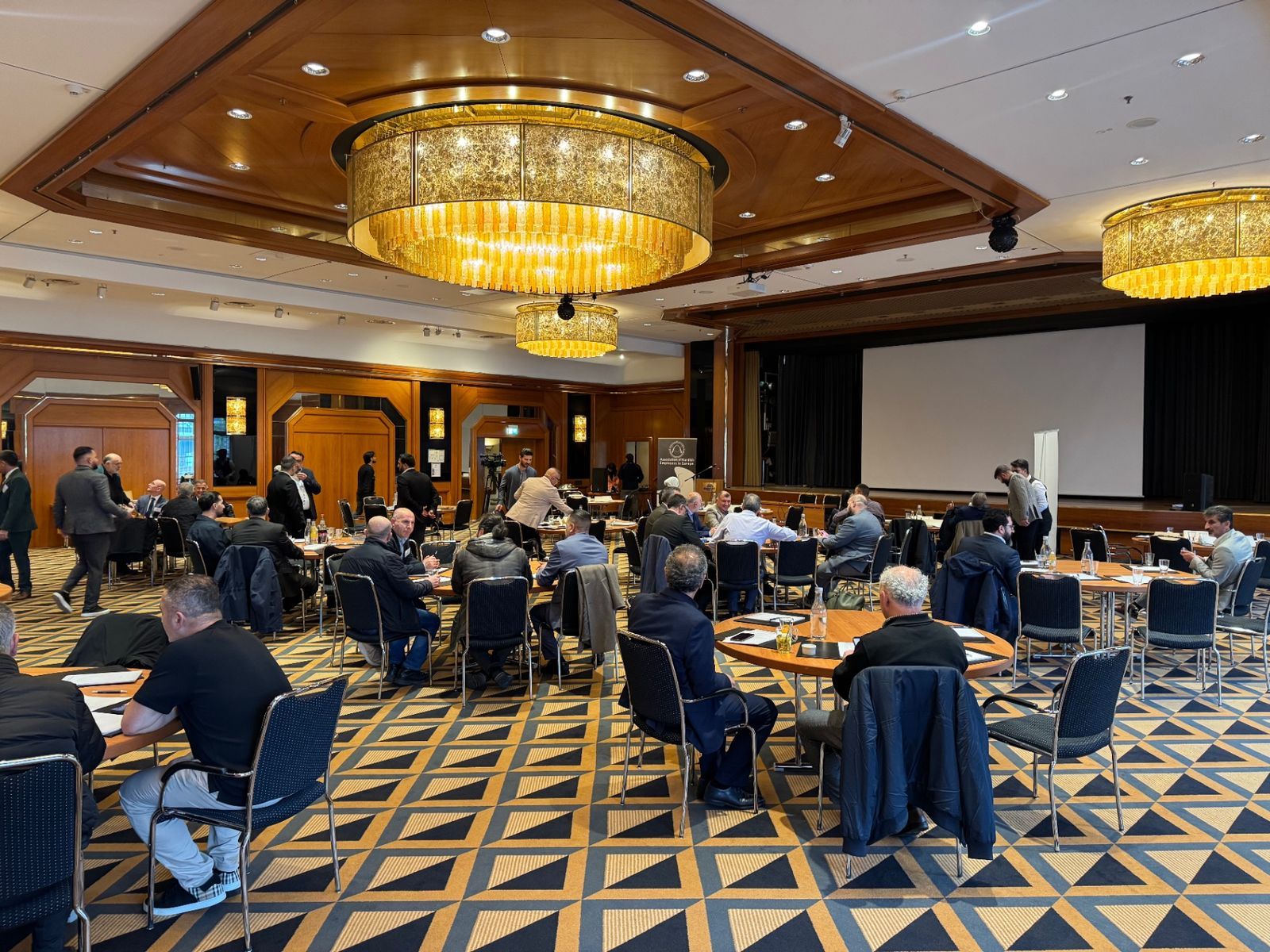 Conference hall of the 11th General Assembly of AKEE in Königswinter: participants at round tables beneath large chandeliers, the stage and screen at the back of the room.