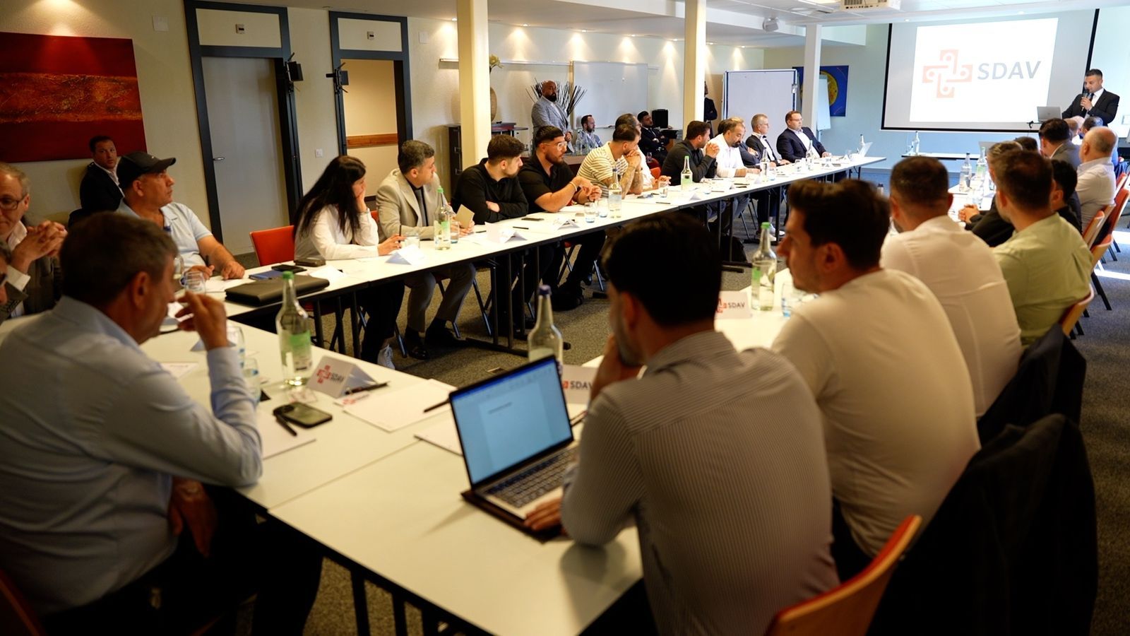 View across the U-shaped table: guests listening to a speaker at the front of the room, SDAV name cards and a laptop on the table.