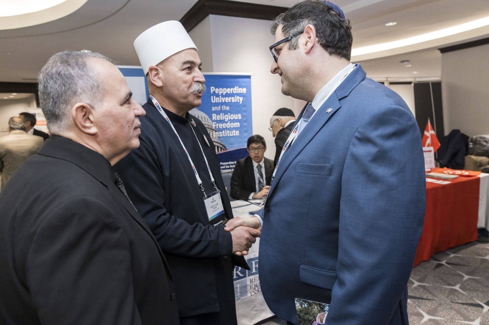 Three participants in conversation in the exhibition area of the summit, two of them shaking hands, next to partner stands.