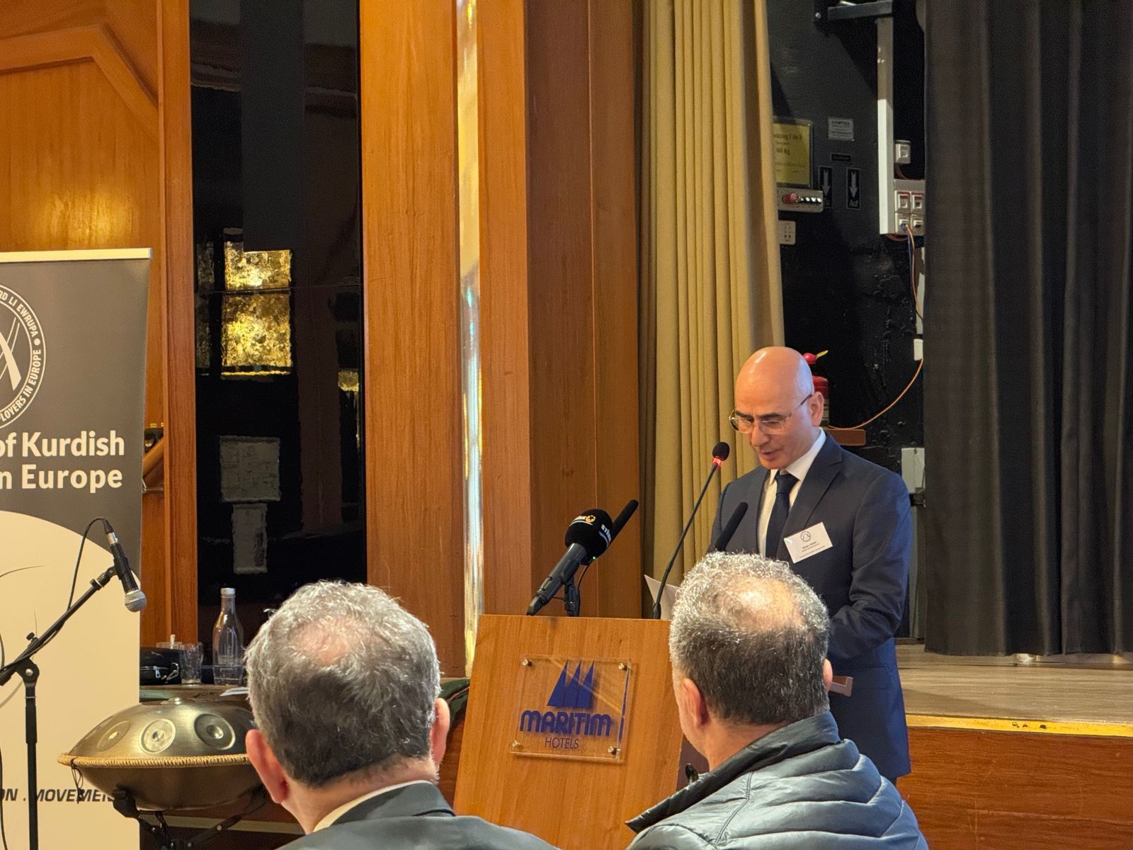 A speaker at the lectern of the AKEE assembly, the AKEE roll-up banner to the left, two listeners in the foreground.