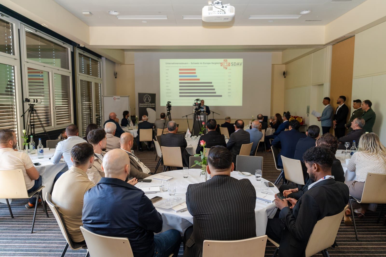 The conference room at the Mövenpick Hotel Egerkingen seen from the back on 26 September 2026: guests at round tables facing the screen and the lectern.