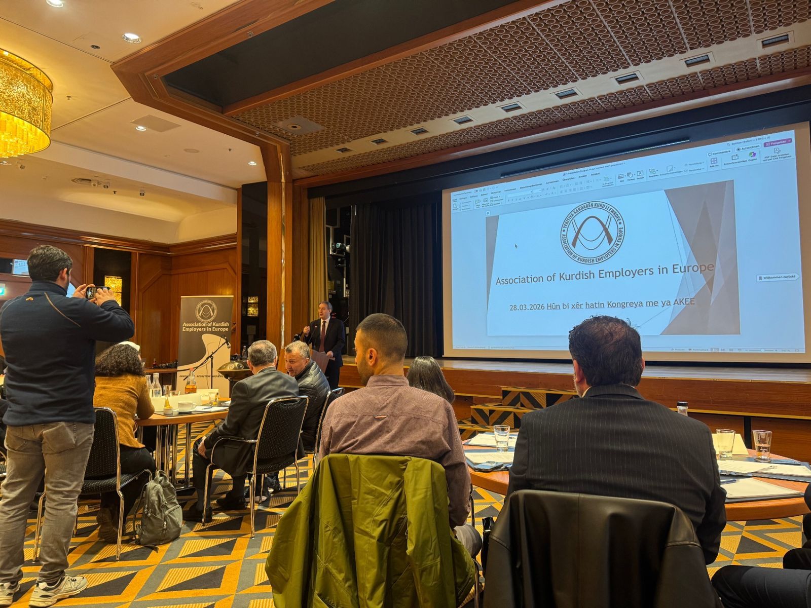 Participants seated at tables in front of the stage; the screen shows the welcome slide of the AKEE assembly with its logo and the date 28.03.2026.
