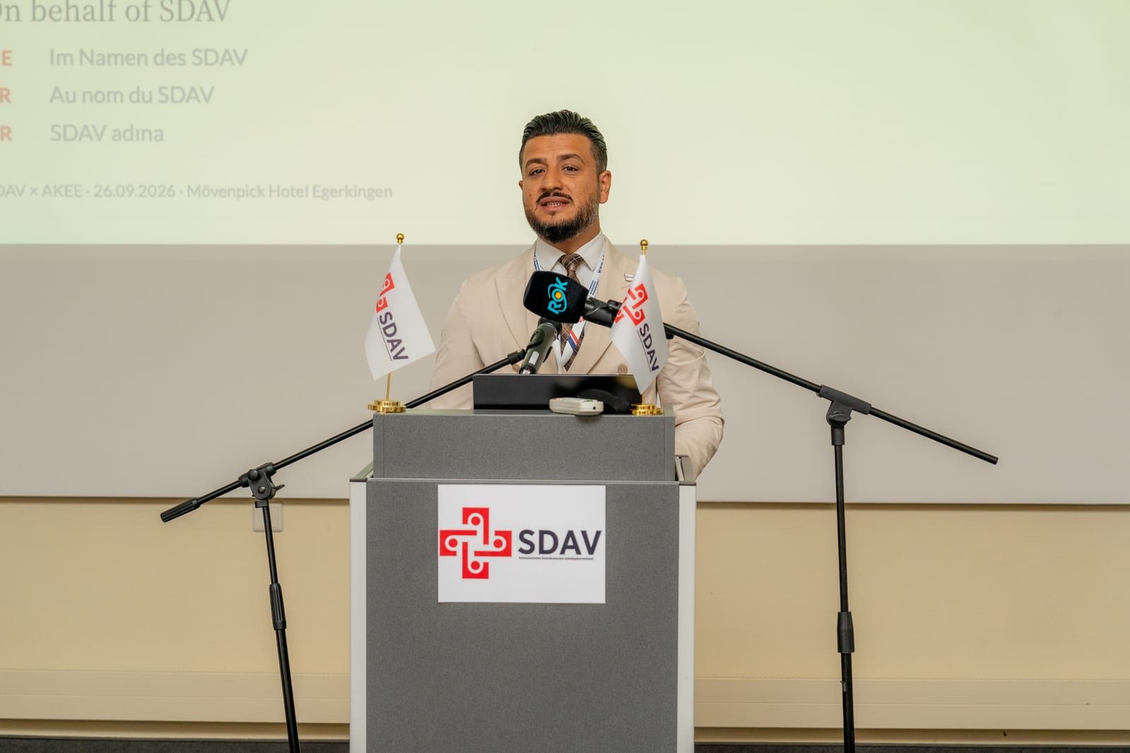 Eyyüp Kavak opening the day at the SDAV lectern; the slide behind him reads “On behalf of SDAV” in four languages.