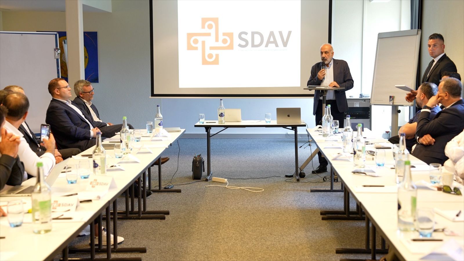 A speaker at the lectern with a microphone in front of the projected SDAV logo, watched by participants on both sides of the room.