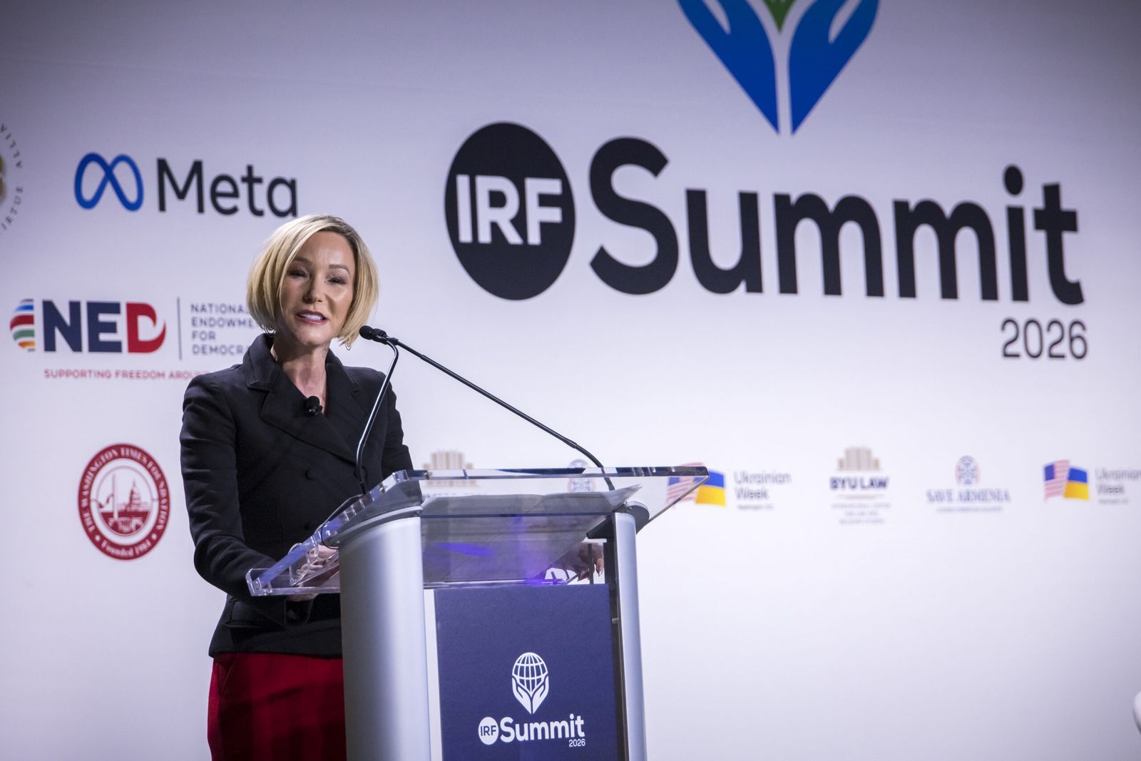 A speaker at the lectern of the main stage, the IRF Summit 2026 backdrop and partner logos behind her.