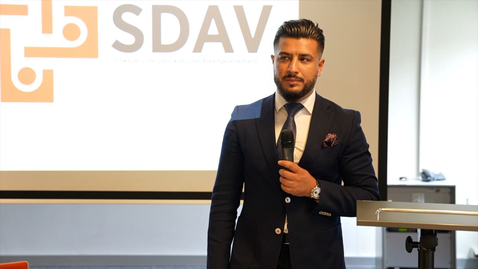 A speaker in a dark suit holding a microphone in front of the projected SDAV logo.