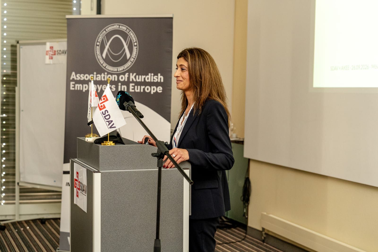 Leylan Uca of AKEE speaking at the lectern, smiling, the AKEE roll-up banner behind her.