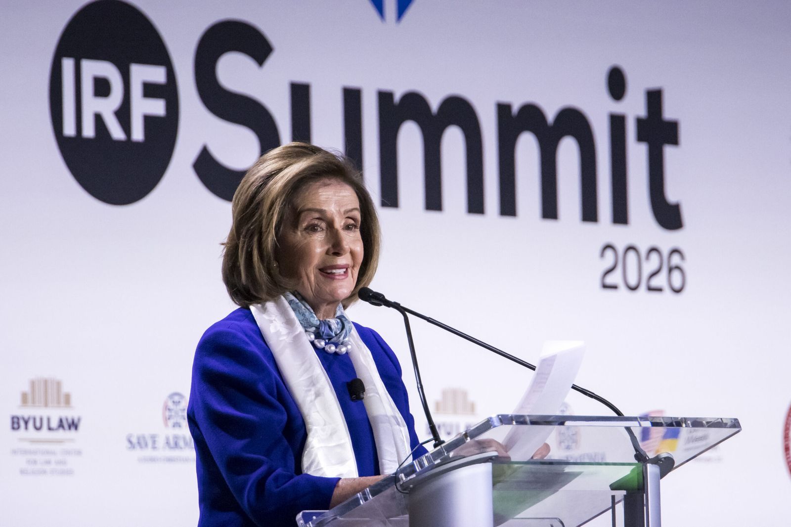 A speaker at the lectern reading from her notes, in front of the IRF Summit 2026 lettering.