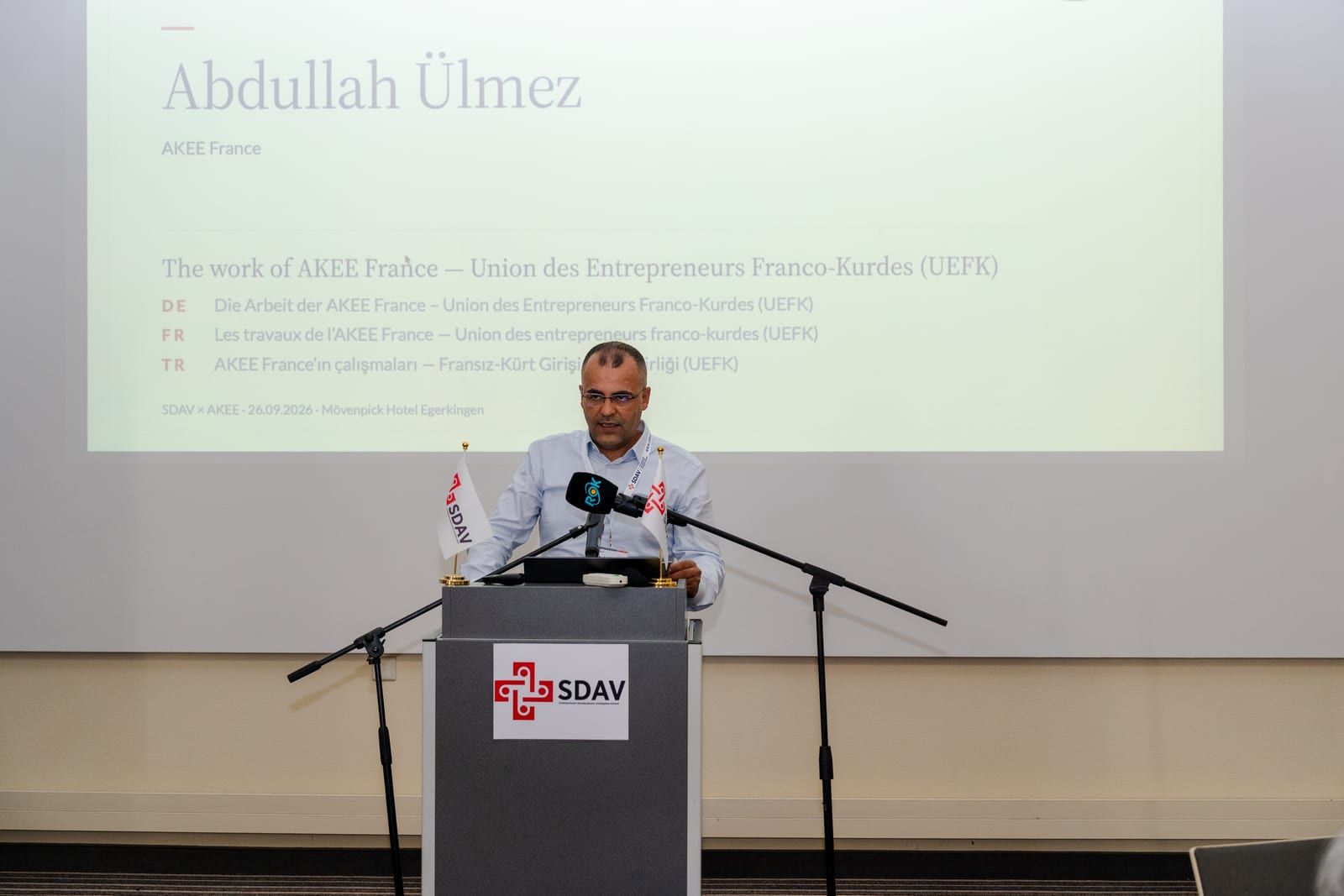 Abdullah Ülmez at the lectern; the slide announces the work of AKEE France – UEFK.