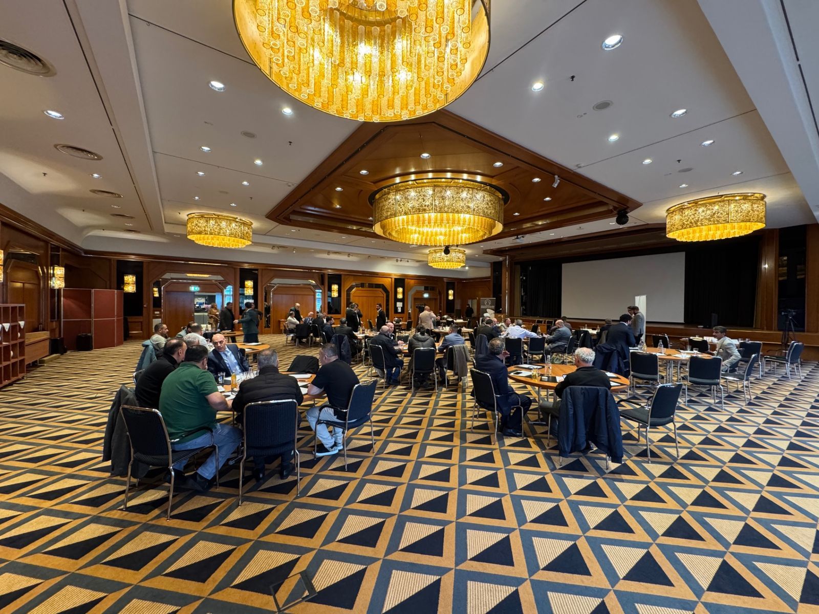 Wide view of the conference hall in Königswinter: participants in conversation at round tables, chandeliers above, the screen at the far end.