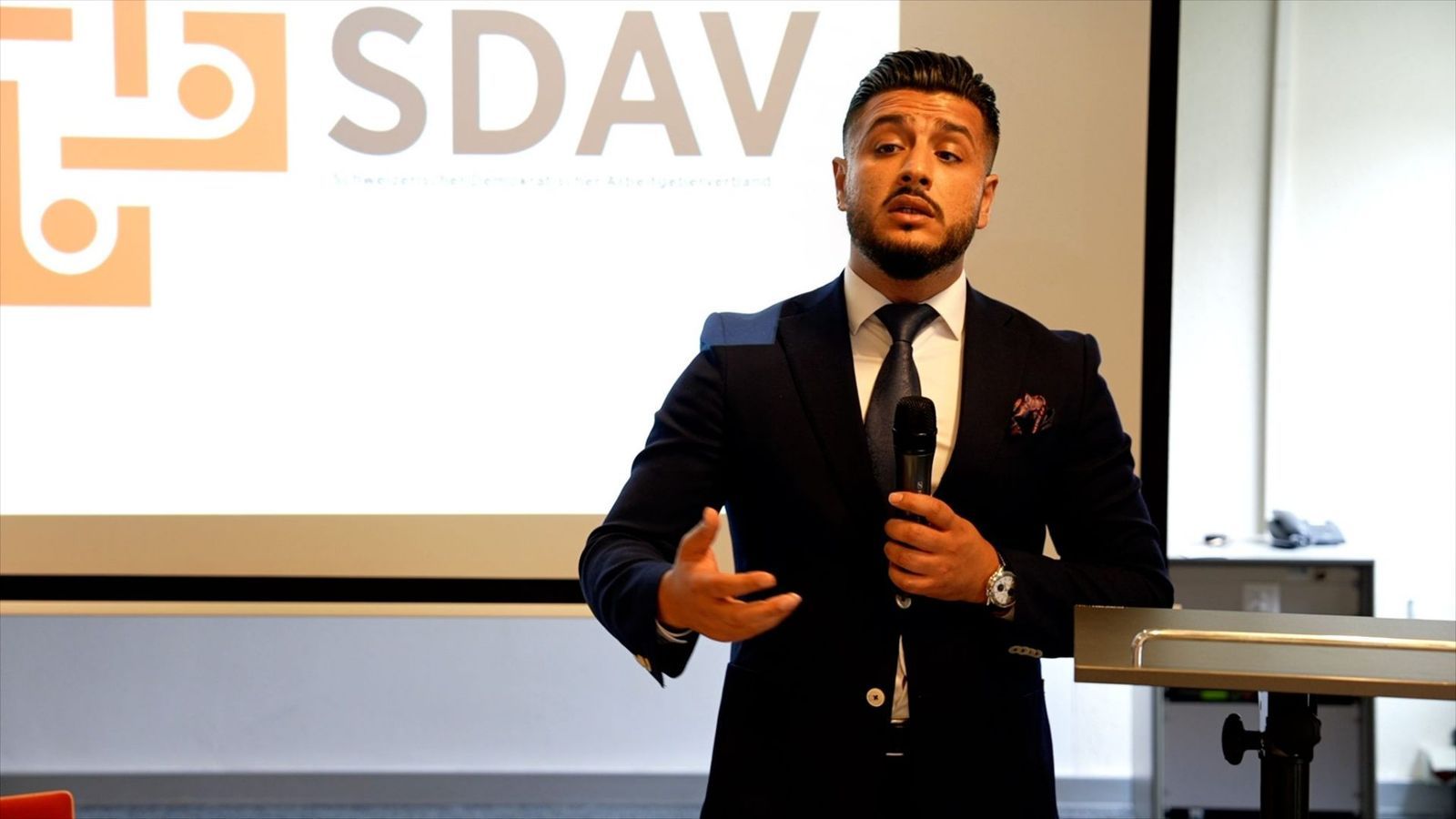 A speaker gesturing as he speaks into a microphone, the SDAV logo on the screen behind him.