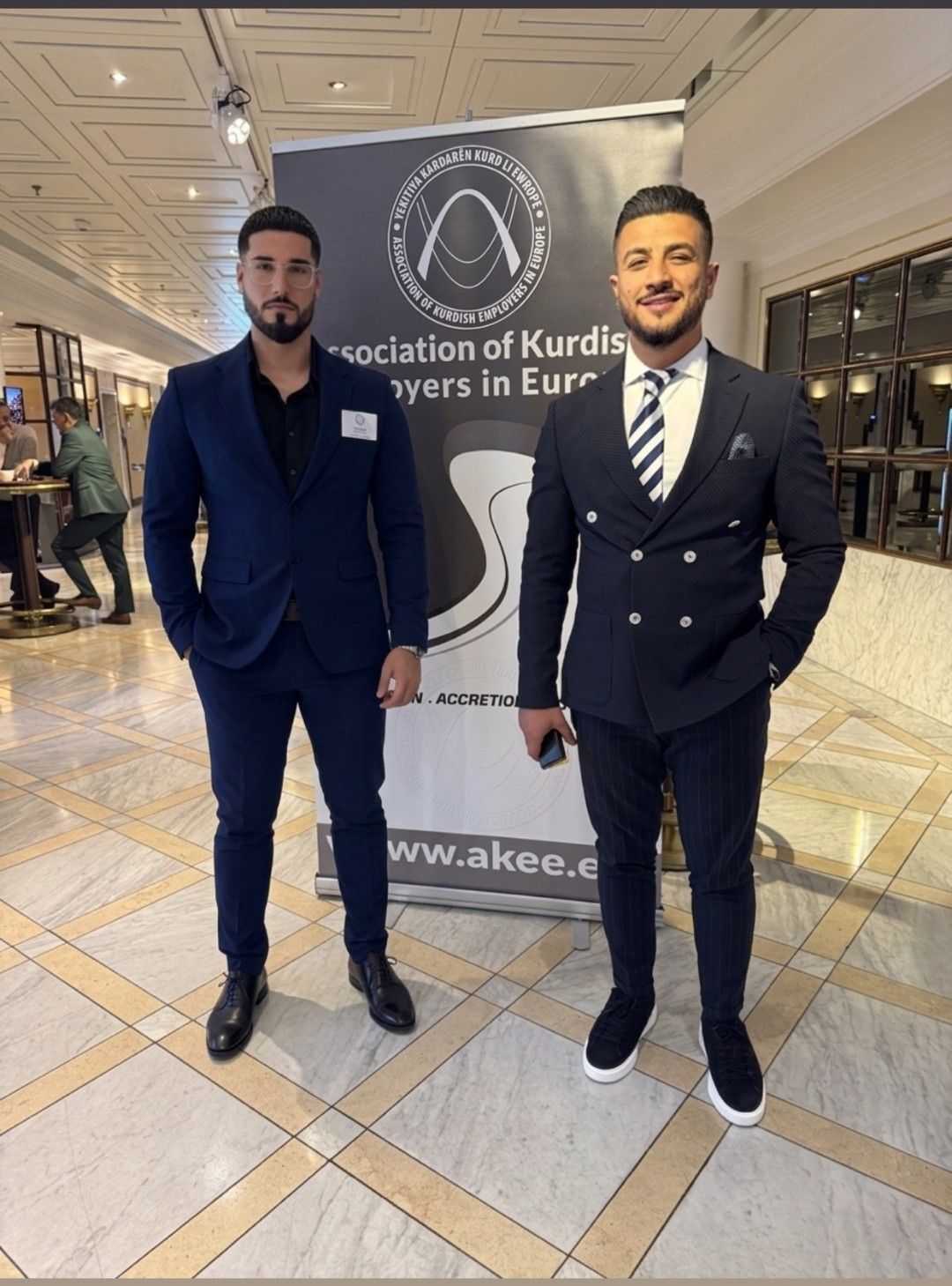 Two members of the SDAV delegation standing in front of the AKEE banner in the hotel foyer in Königswinter.