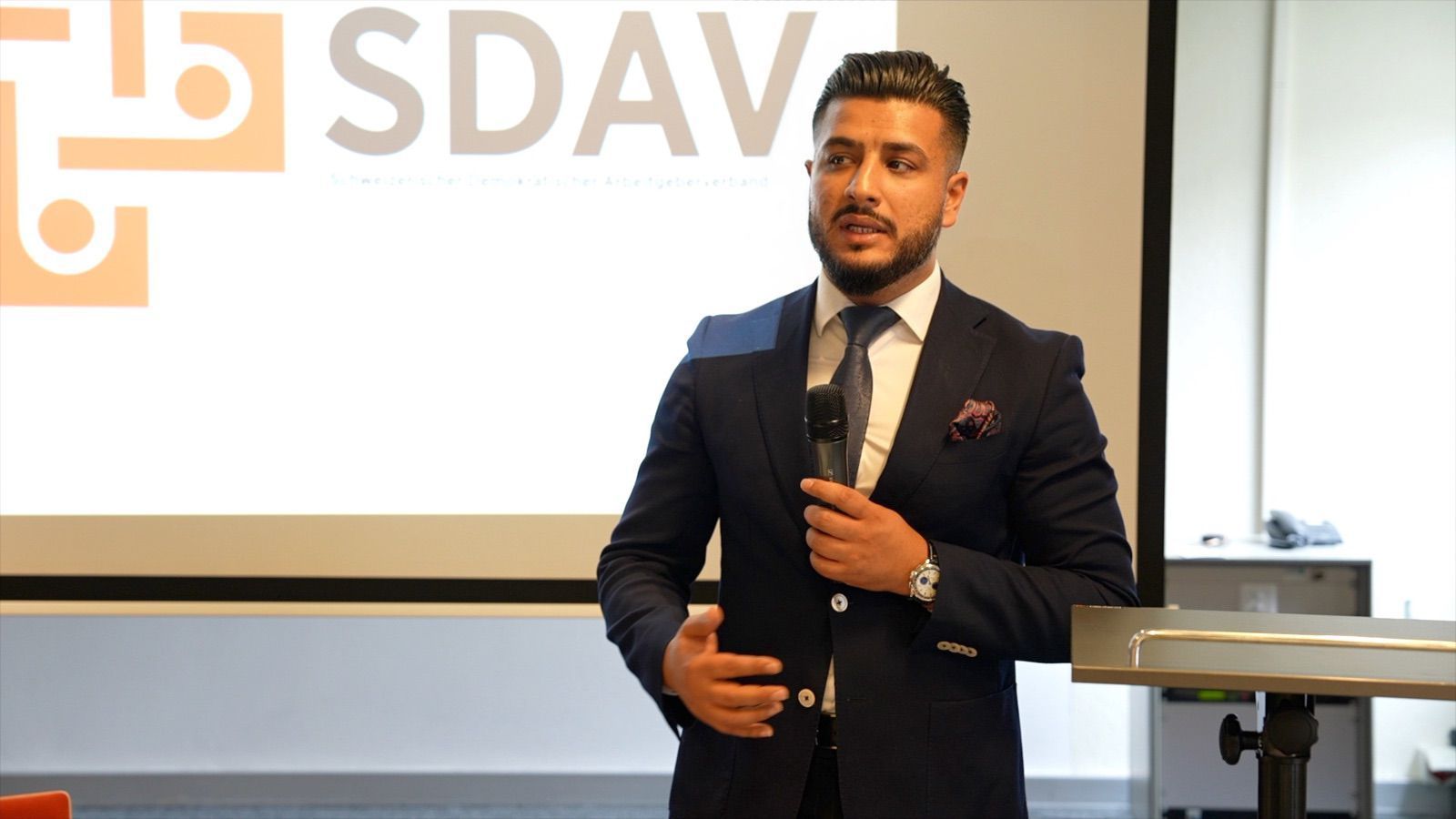 Close view of a speaker with a microphone in front of the SDAV logo.