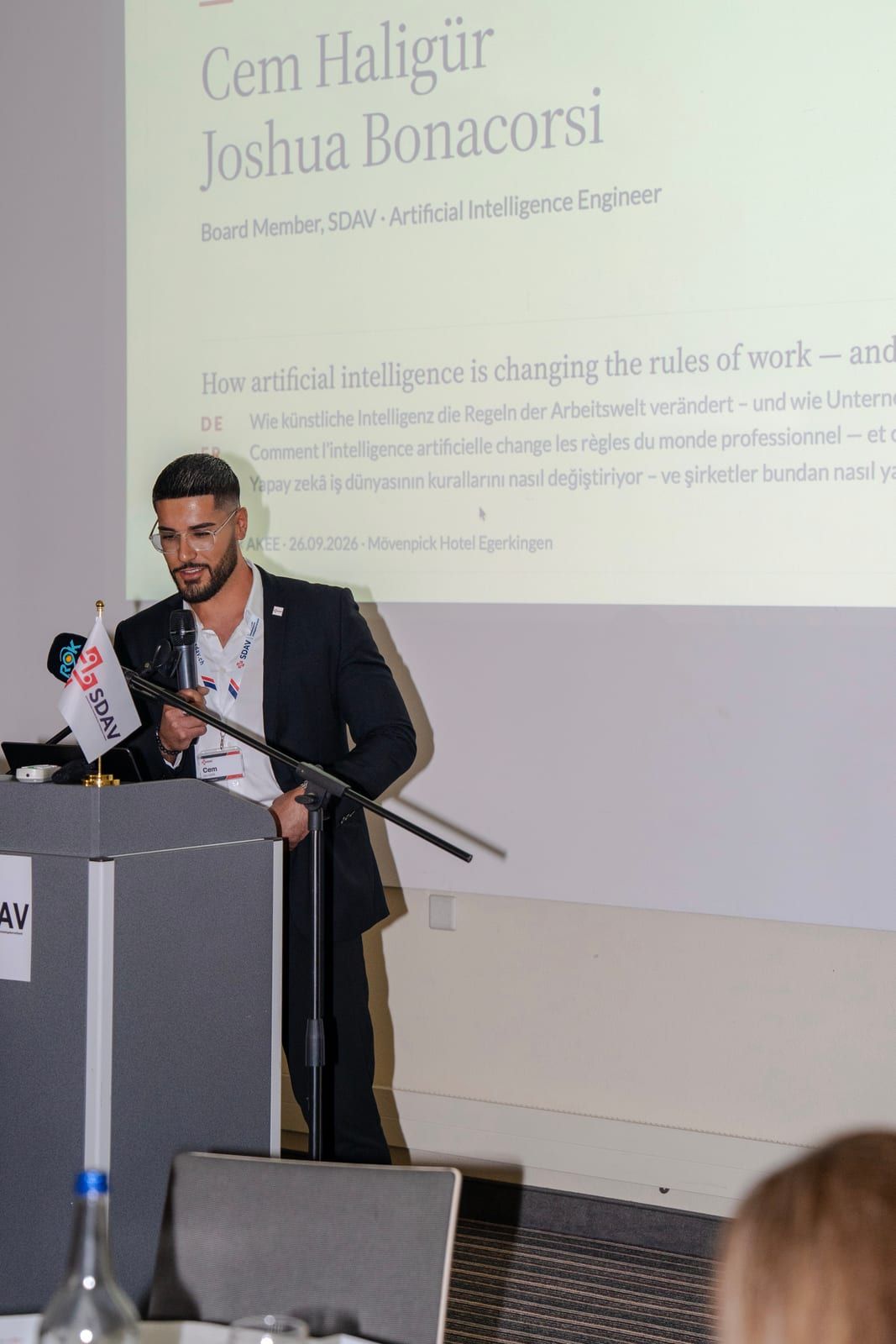Cem Haligür speaking with a microphone, the title slide of the talk on artificial intelligence projected behind him.