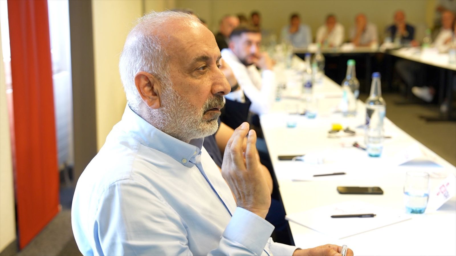 A participant, seen in profile, making a point during the discussion, other guests along the table in the background.