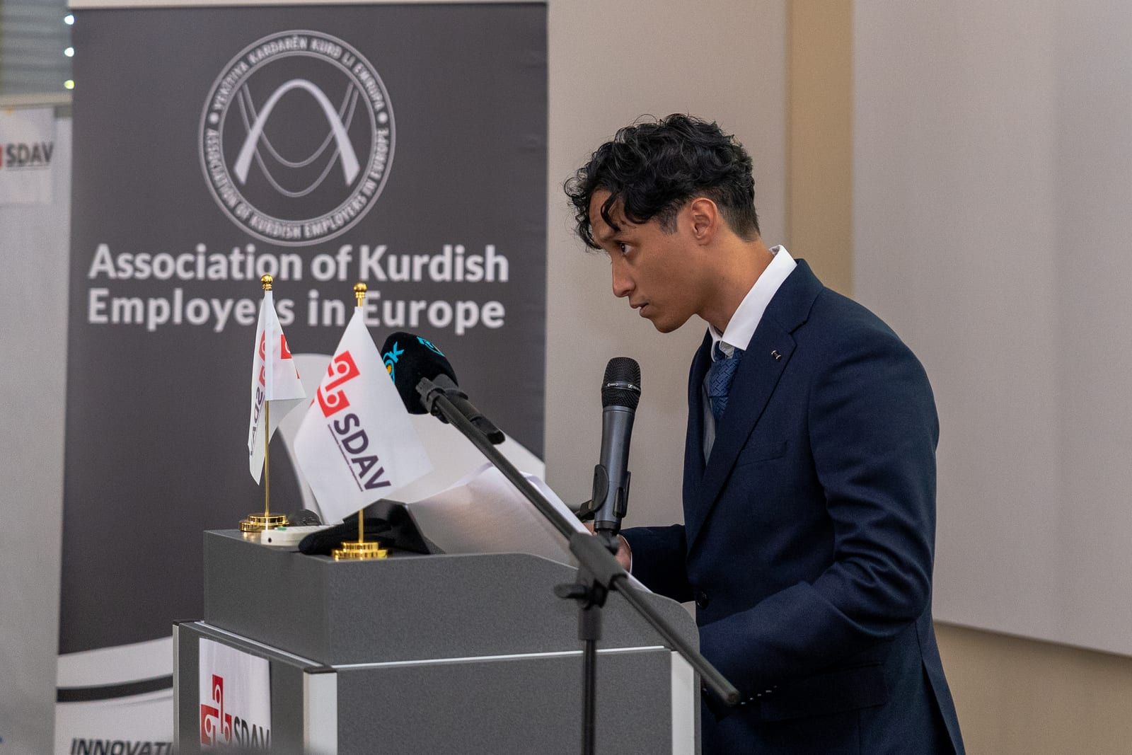 Joshua Bonacorsi at the lectern in profile, SDAV table flags and the AKEE banner beside him.
