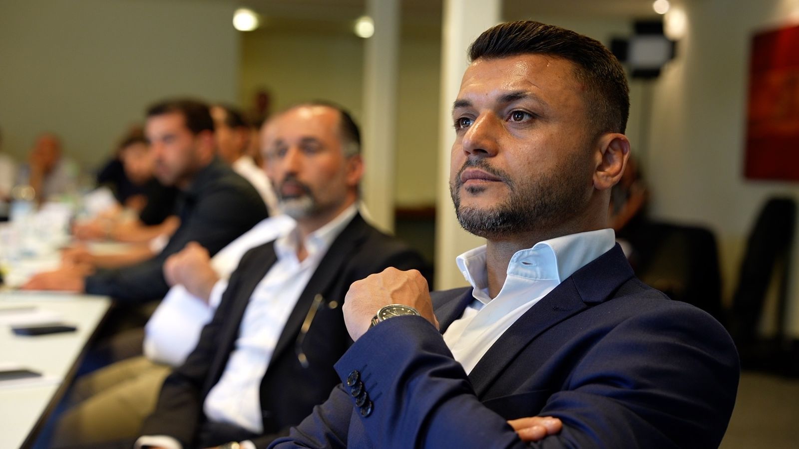 A participant listening attentively, arms crossed, other guests along the table behind him.