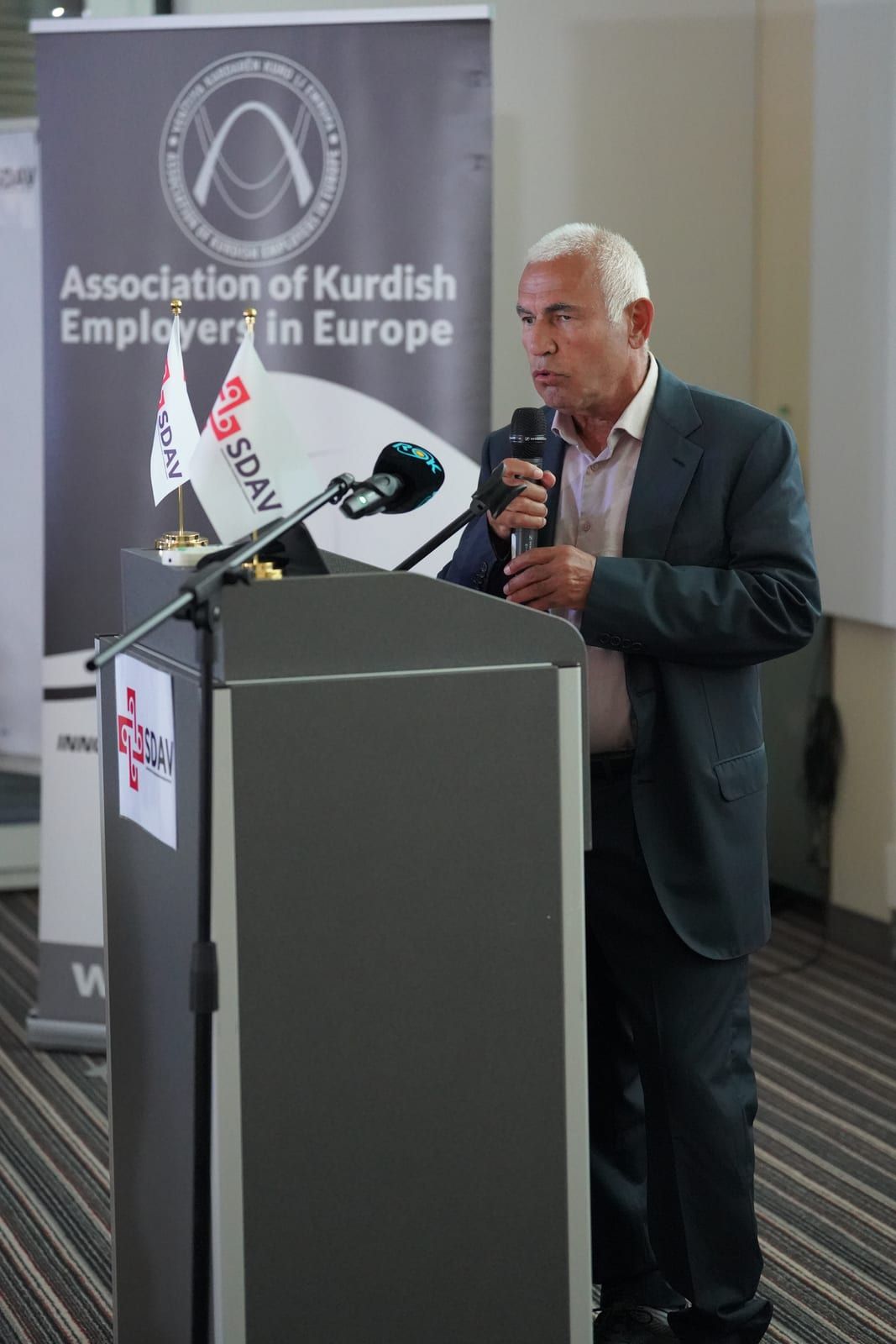 Sobhi Aksoy speaking with a handheld microphone in front of the AKEE roll-up banner.