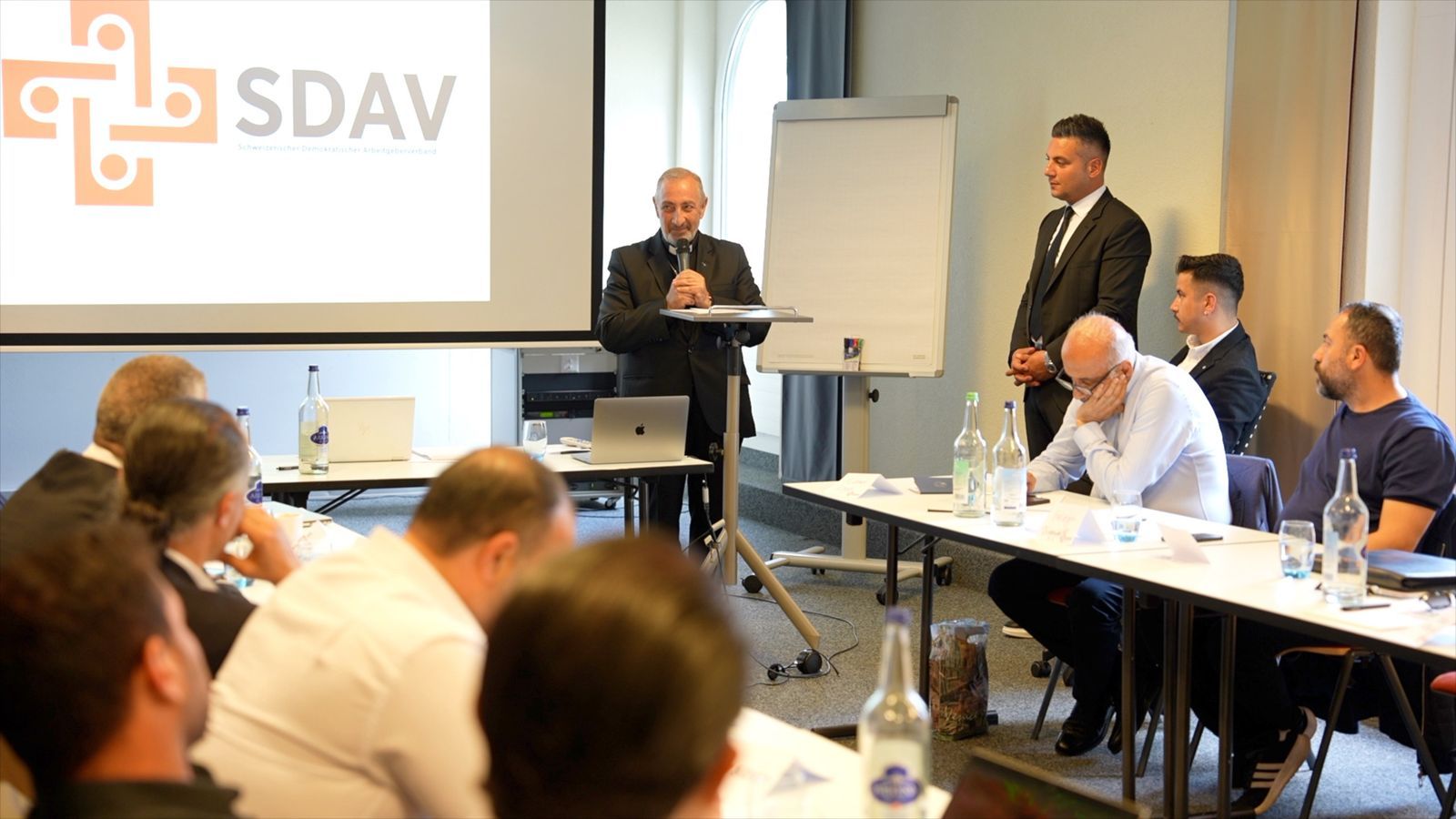 A speaker at the lectern with a microphone next to a flip chart, in front of the SDAV logo, participants listening on both sides.