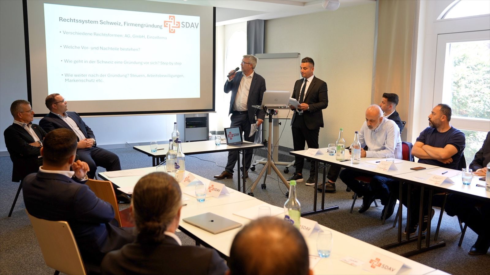 Two speakers presenting in front of a slide on the Swiss legal system and company formation, participants around the U-shaped table.