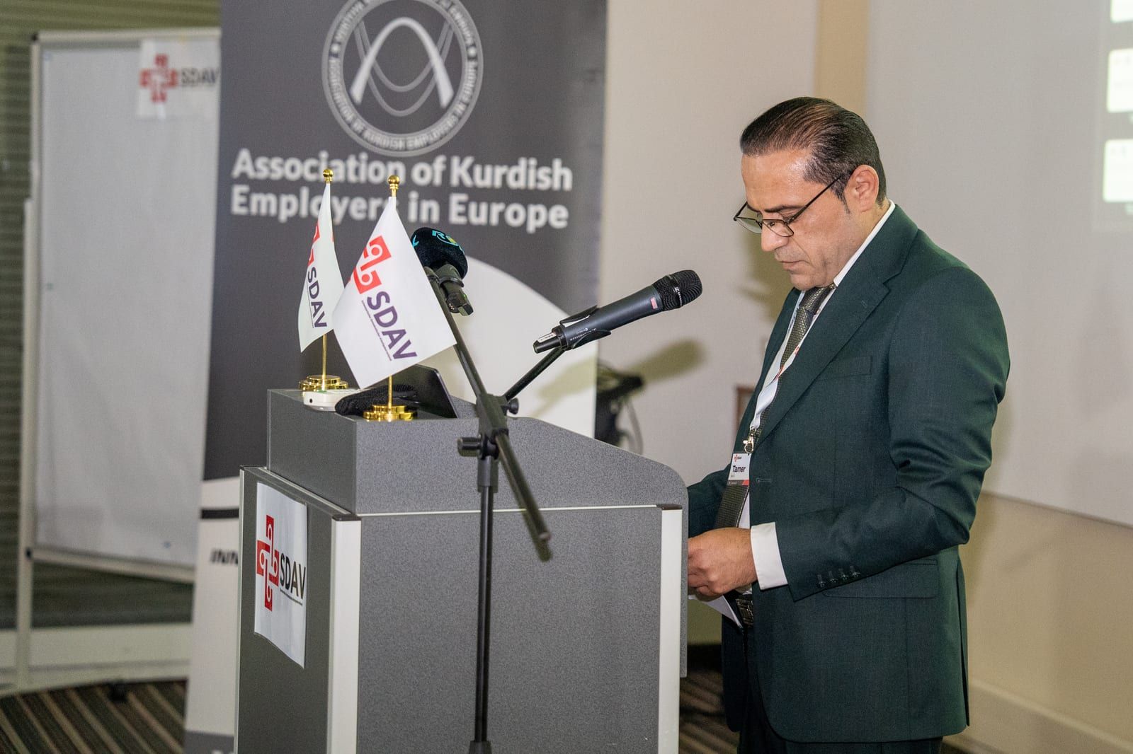 Tamer Guli at the lectern reading from his notes, the AKEE banner behind him.