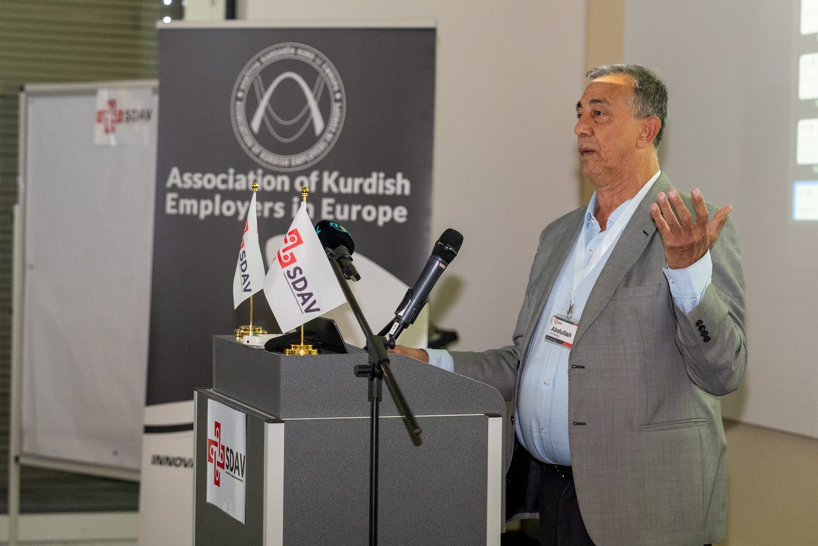 Abdullah Demirbaş speaking at the lectern with an open hand, the AKEE banner behind him.