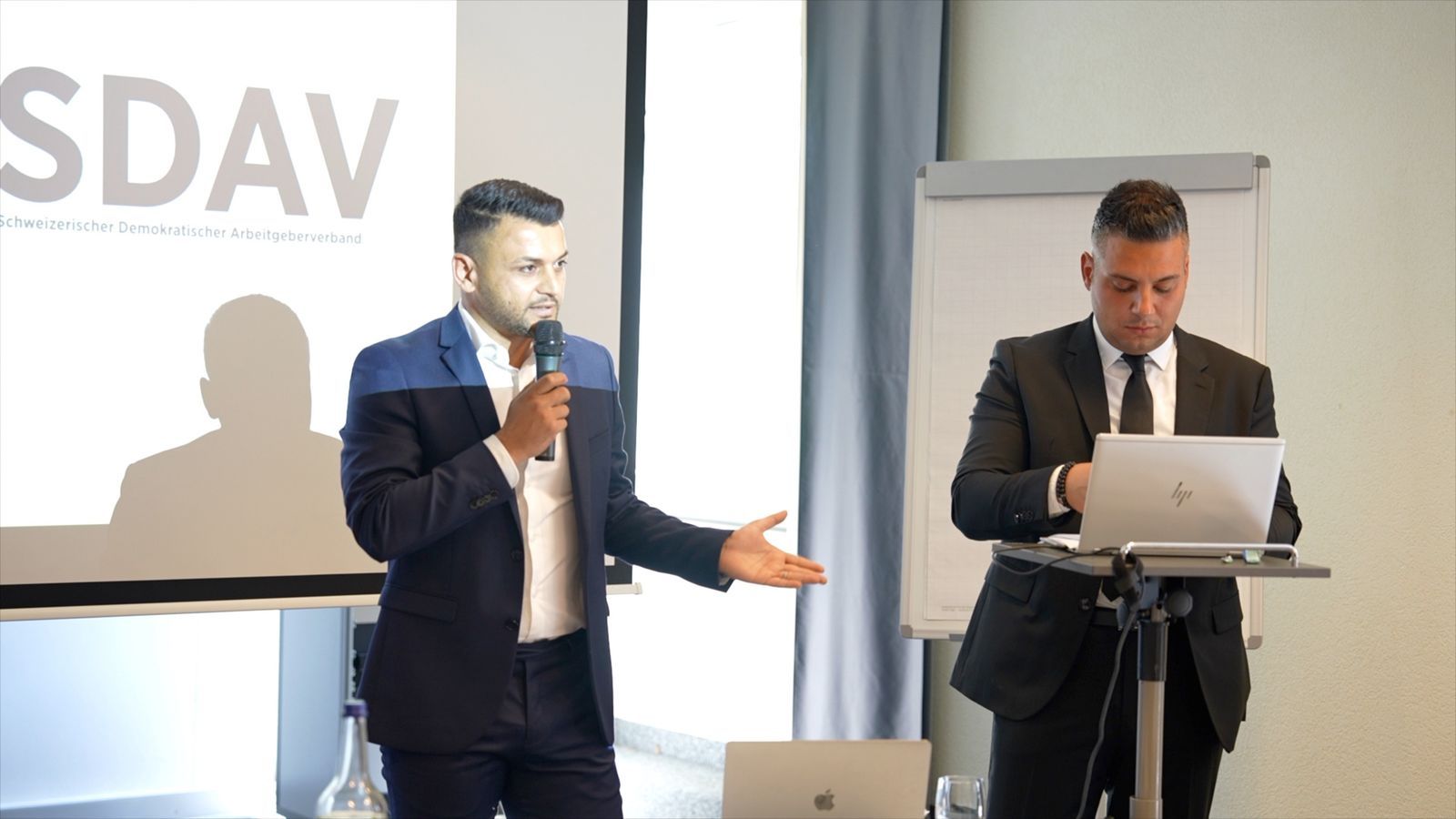 Two speakers at the front of the room: one speaking into a microphone, the other at a laptop on the lectern, in front of the SDAV lettering.