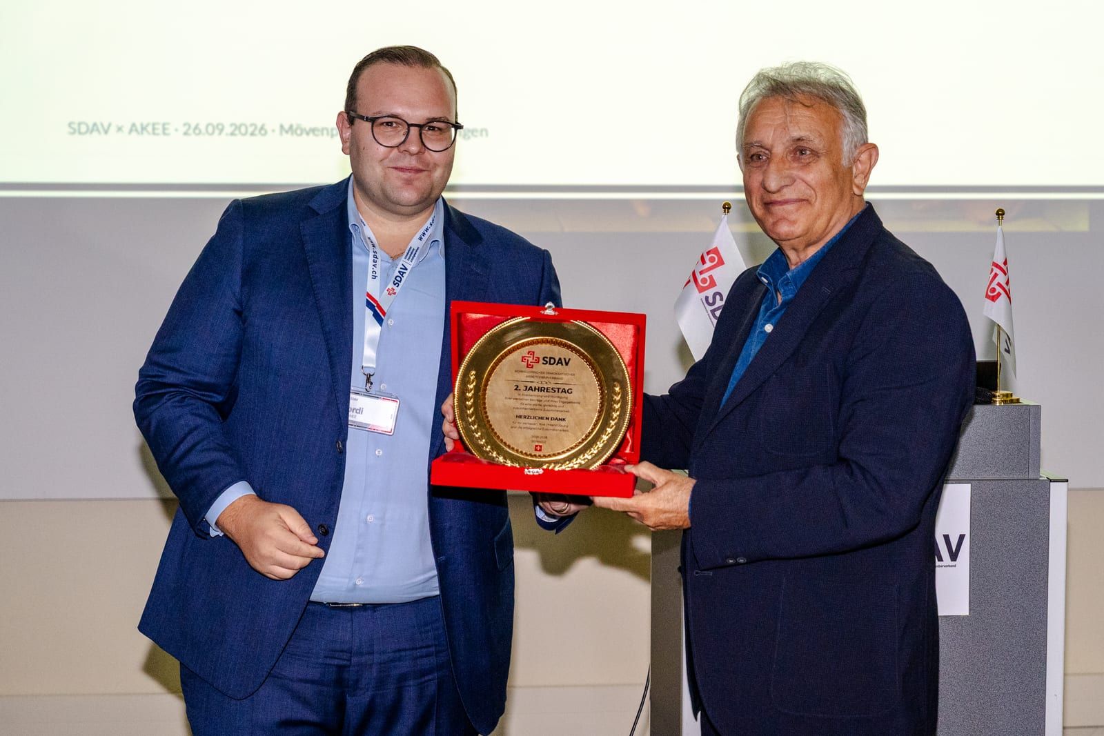 Jordi Alvarez and Erol Katırcıoğlu holding an anniversary medallion in its red box, both smiling at the camera.