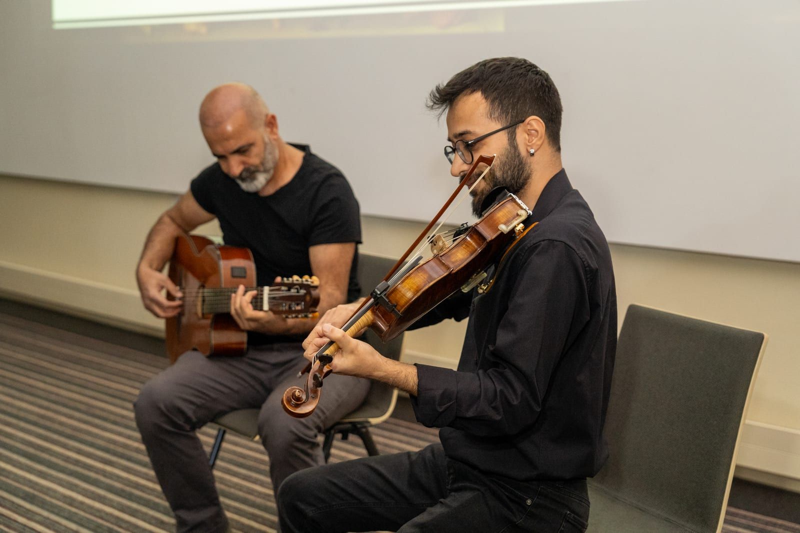 A guitarist and a violinist playing seated side by side, the violinist in the foreground with his bow raised.