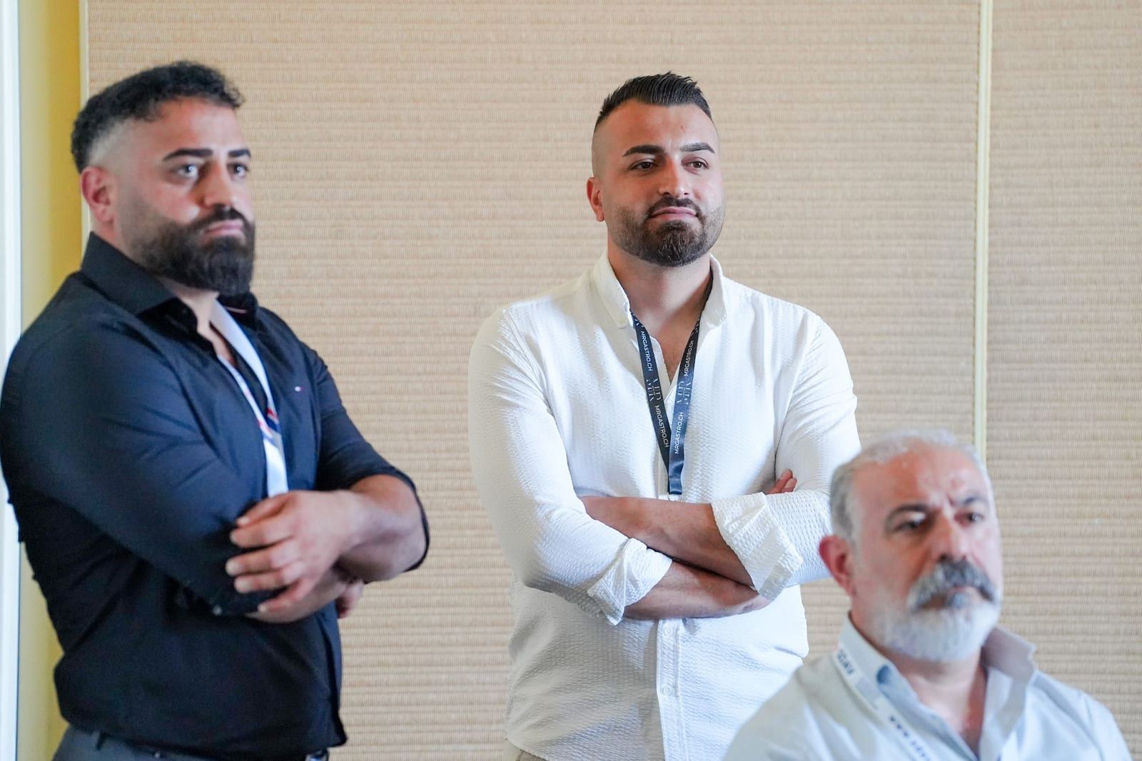 Two participants standing against the wall listen to a speaker, arms crossed.