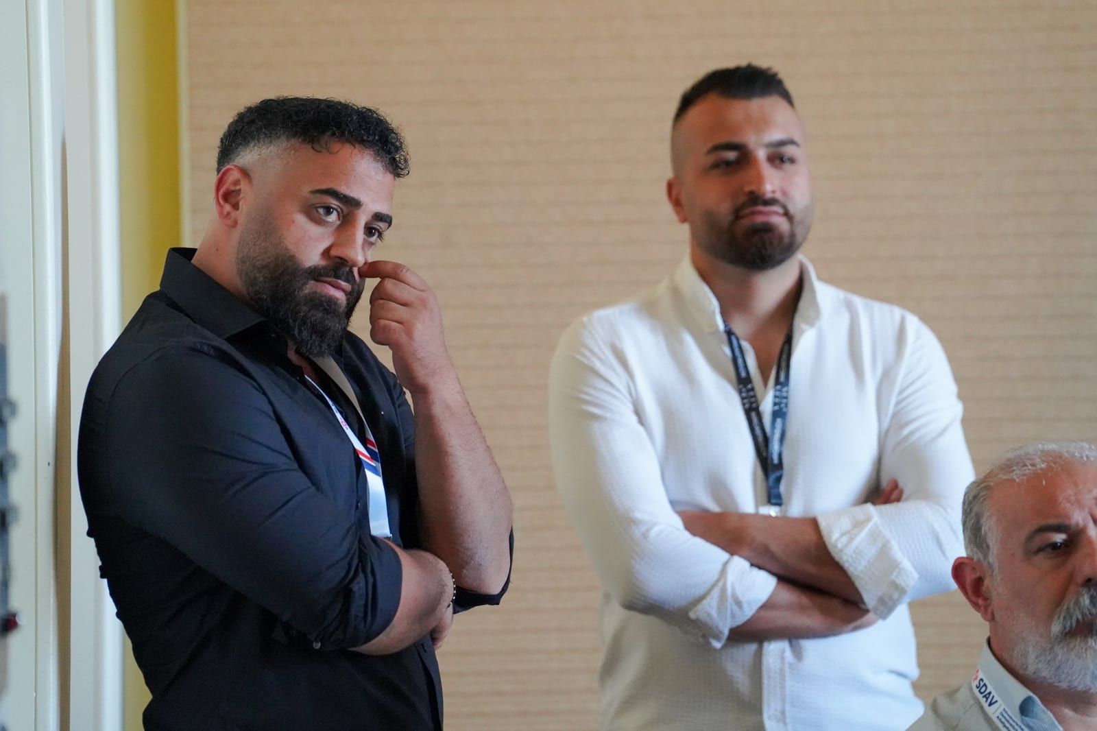 A participant in a black shirt listens, hand to his face, next to a second participant standing at the back of the room.